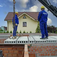 Dwóch budowlańców w niebieskich kombinezonach montuje pustaki stropowe. Dźwig podnosi kolejne elementy.