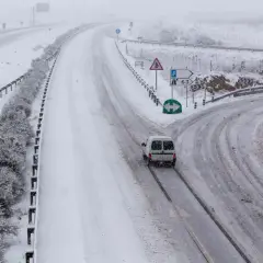 Nieve en la sierra de Madrid hoy. Una furgoneta blanca circula por una carretera nevada, con señales de tráfico y vegetación cubierta de blanco.