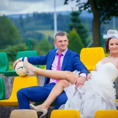 Niespodzianka dla pary młodej: pan młody w garniturze i panna młoda w sukni ślubnej siedzą na trybunach, ona opiera nogę na piłce.