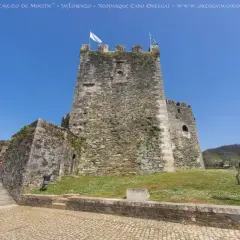El castillo de Moeche, una imponente fortaleza de piedra, se alza bajo un cielo azul. Un puente de madera conduce a sus muros cubiertos de vegetación.