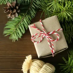 Świąteczne zestawy dla rodziny: prezent owinięty w papier, gałązki choinki, szyszki i czapka z pomponem na drewnianym tle.