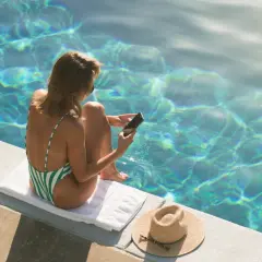 Kobieta w zielono-białym stroju kąpielowym siedzi przy basenie, trzymając telefon. Nie rób tego, kiedy on nie odpisuje, bo słońce i woda kuszą.