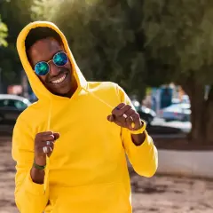 Un homme souriant porte un sweat jaune vif, ses lunettes rondes reflétant le soleil. Le grammage du sweat semble parfait pour une journée ensoleillée.