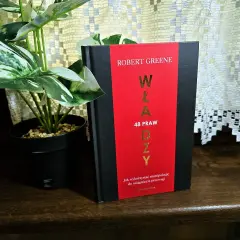 Książka "48 praw władzy" Roberta Greene'a na drewnianej półce, obok rośliny w czarnej doniczce.