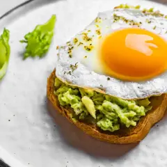Un toast garni d'avocat écrasé et surmonté d'œufs sur le plat, assaisonné d'herbes. Deux feuilles de laitue verte décorent l'assiette.