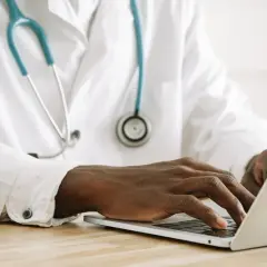 Mani di un medico che digita su un laptop, con uno stetoscopio appeso al camice bianco. Forse sta gestendo gli arretrati medici di base quando.