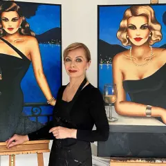 Künstlerin steht zwischen zwei Gemälden von eleganten Frauen in schwarzen Kleidern mit Gläsern Wein.