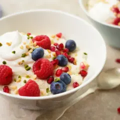 Veganes Grießbrei-Dessert mit frischen Himbeeren, Blaubeeren, Granatapfelkernen und gehackten Nüssen, garniert mit einem Klecks veganer Sahne.