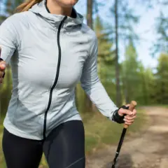 Kobieta w szarej kurtce i czarnych legginsach uprawia north walking na leśnej ścieżce, ciesząc się aktywnością na świeżym powietrzu.