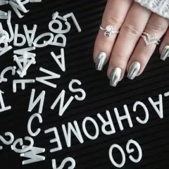 A hand with silver metallic nails and rings rests on a black surface scattered with white letters.