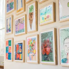 A vibrant kids art gallery wall showcases colorful drawings of ice cream, flowers, and portraits, each framed in light wood.