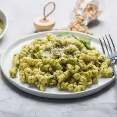 Pyszny makaron z pesto ze słoika, posypany parmezanem. Obok miseczka z zielonym sosem i kawałki sera.