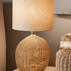 Lámpara de mesa mimbre con pantalla de tela, junto a una pila de libros sobre una cesta de mimbre.