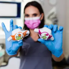 Dentystka w niebieskich rękawiczkach prezentuje dwa opakowania z zabawnymi dinozaurami, sugerujące zastosowanie w leczeniu, być może z użyciem podtlenku azotu.