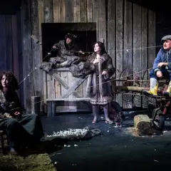 Scena z życia wiejskiego w sztuce teatralnej. Cztery postacie w rustykalnych strojach, na tle drewnianej stodoły.