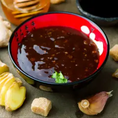 Sos teriyaki przepis: miseczka z ciemnym sosem, obok plasterki imbiru, czosnek, cukier i miód.