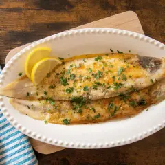 Due filetti di sogliola alla parmigiana dorati, guarniti con prezzemolo tritato e spicchi di limone, serviti su un piatto bianco.