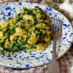 Un delicioso revuelto de espinacas cremoso, servido en un plato con salpicaduras azules. Acompañado de pan, ajo y huevos frescos.