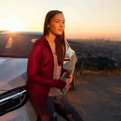 Ung kvinna lutar sig mot en vit Mercedes med en flaska i handen, vid solnedgången över en stad.