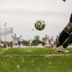 Piłkarz kopie piłkę na boisku, w tle inni zawodnicy i bramka.