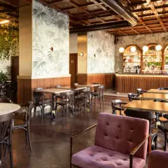 Interno di un locale con carta da parati tropicale, bancone bar illuminato e tavoli in legno. Perfetto per scoprire i **cheap bars in Milan**.