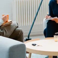 Dwie osoby siedzą w pokojach, jedna pije kawę, druga pisze.