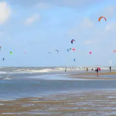Plaża z wieloma latawcami do kitesurfingu na niebie i ludźmi spacerującymi po piasku.