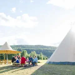 En familj njuter av en solig dag på alevi camping i Sverige. De sitter runt en lägereld under ett stort parasoll, med en tipi i bakgrunden.