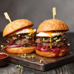 Dwa soczyste burgery z jajkiem sadzonym i dodatkami na desce, obok miseczka sosu.