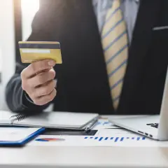 Biznesmen trzyma kartę kredytową, planując wykorzystać kredyt odnawialny do rozwoju firmy. Na biurku laptop i wykresy.