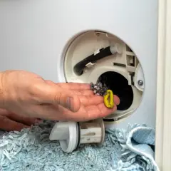 Brudny filtr w pralce, pełen kłaczków i włosów, trzymany w dłoni. Obok leży wyjęty filtr.