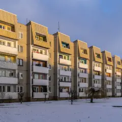 Długa, szara wielka płyta z balkonami i charakterystycznymi, skośnymi dachami, oświetlona słońcem. Na śniegu widać ślady.