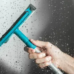 Main tenant une raclette bleue pour le nettoyage vitre salle de bain, laissant des traces d'eau sur la paroi.