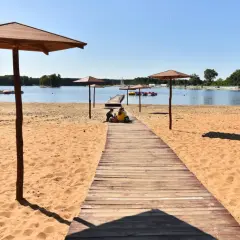 Sielpia jezioro: drewniana ścieżka prowadzi na piaszczystą plażę pod parasolami. W oddali widać pomost z rowerami wodnymi.