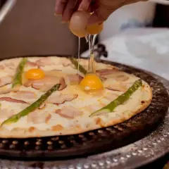 Ręka wylewa jajko na pizzę z boczkiem i szparagami. Pyszna pizza z jajkiem w przygotowaniu.