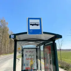 Szklane przystanki autobusowe z tabliczką informującą o transporcie publicznym, otoczone drzewami i zielonym polem.