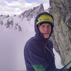 Alpinista con casco amarillo y ropa de montaña, rodeado de nieve y niebla en un paisaje rocoso.