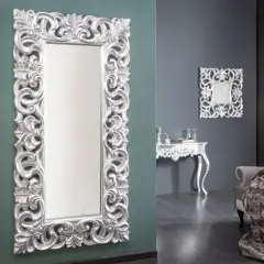 Eleganckie, wysokie lustro w srebrnej ramie z bogatymi zdobieniami, stojące obok wazonu z gałązkami.