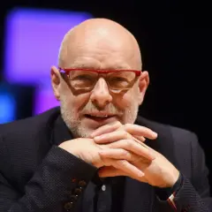 Brian Eno, eno musicista inglese, con occhiali rossi e mani intrecciate.