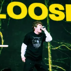 Wokalista z otwartymi ustami śpiewa do mikrofonu na tle sceny z napisem "LOOSE".
