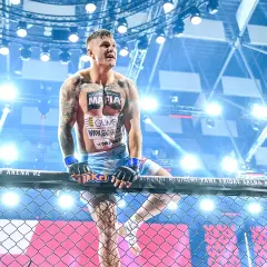 Alan Kwieciński, wytatuowany zawodnik MMA, w rękawicach, oparty o siatkę klatki, w świetle reflektorów.