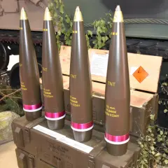 Cztery pociski amunicji artyleryjskiej 155 mm, każdy z oznaczeniem TNT, stoją obok drewnianych skrzyń i gąsienic czołgu.