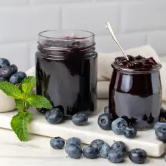 Due barattoli di marmellata di mirtilli, uno aperto con un cucchiaino, circondati da mirtilli freschi e foglie di menta.