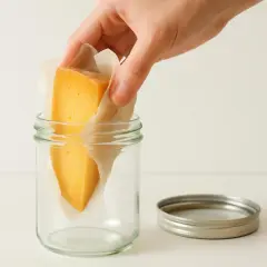 A hand places a wedge of cheese wrapped in parchment paper into a glass jar.