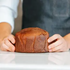 Manos sostienen un pan brioche dorado y esponjoso sobre una superficie blanca.