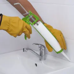 Hände mit Handschuhen dichten mit einer Kartusche den Rand eines Waschbeckens ab. Frage: wie lange muss silikon trocknen dusche?