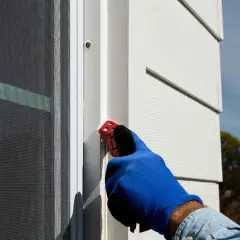 A gloved hand uses a red tool to smooth caulk along a window frame, demonstrating how to caulk windows for a weather-tight seal.