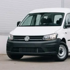 Biały Volkswagen Caddy, samochód osobowy zarejestrowany jako ciężarowy bez kratki, stoi przed szarą ścianą.