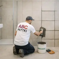 Fachowiec w koszulce "ABC PŁYTKI" wykonuje wymianę fugi w płytkach łazienkowych, używając kielni. Obok stoi wiadro z zaprawą.