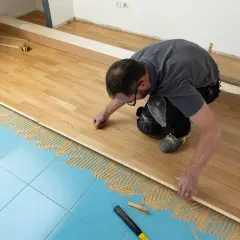 Pose d'un parquet stratifié sur un sol bleu carrelé, avec de la colle appliquée. L'homme ajuste une lame de bois.